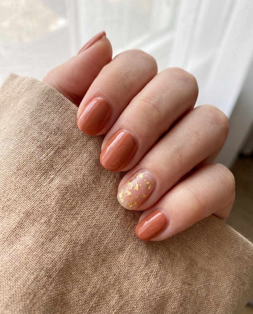 Manicure in warme terracotta tint met een gouden glitter-accentnagel, close-up bij het raam